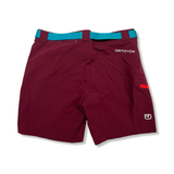 Baksidan av ett par lila vandringsshorts från Ortovox för dam med blått skärp, visade mot en vit bakgrund.