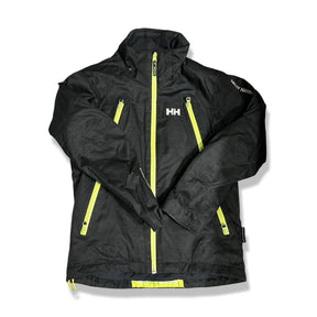 Svart skidjacka från Helly Hansen för barn i storlek 152, med gula dragkedjor, fotograferad framifrån mot en vit bakgrund.