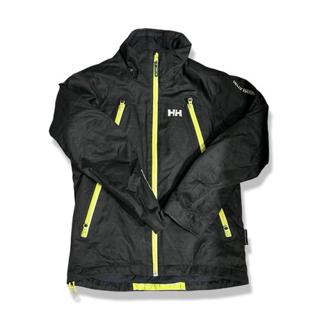 Svart skidjacka från Helly Hansen för barn i storlek 152, med gula dragkedjor, fotograferad framifrån mot en vit bakgrund.
